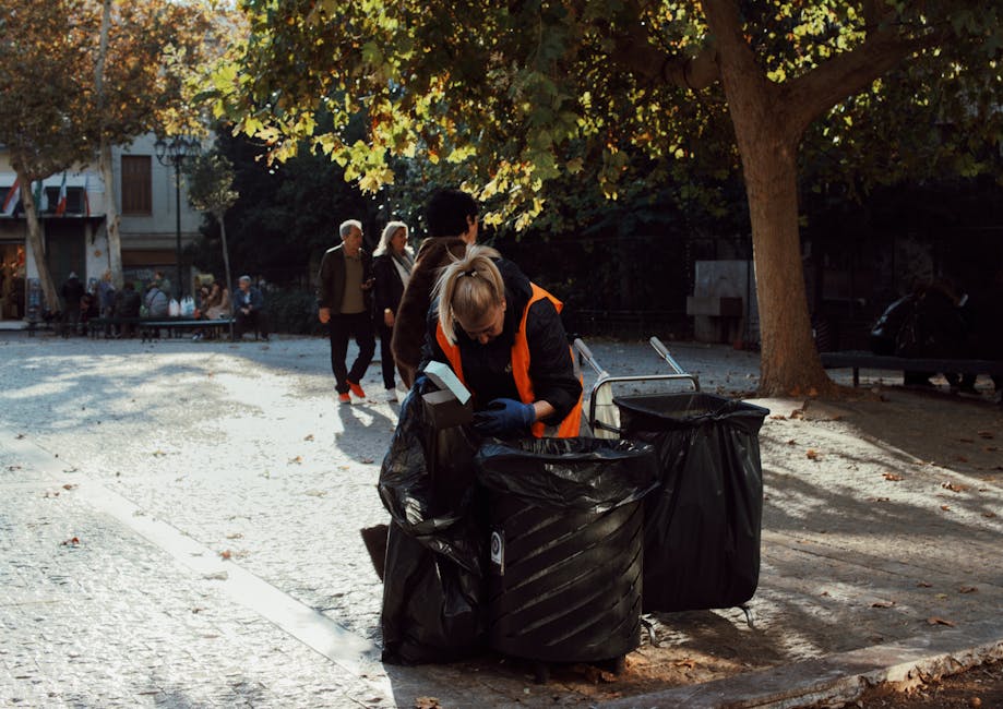 Three individuals dressed in bright orange work uniforms are walking along a paved area beside a calm river or canal. The person in the foreground, a woman with dark skin and a bun hairstyle, is carrying two large blue plastic garbage bags filled with waste in each hand. To her left, a woman with long dark hair tied back is holding a single blue bag, and to her right, a man with light skin and short hair is carrying another blue bag, with an additional larger blue bag slung over his shoulder. The bags appear to contain rubbish, possibly recyclable materials or general waste. In the background, a concrete bridge stretches across the water, and a few trees are visible behind it, indicating a suburban or urban park setting. The scene is lit with natural daylight, with a soft sky suggesting either early morning or late afternoon. This image visually supports concepts of independent waste collection, rubbish clearance, or private waste disposal services, aligning with themes of rubbish removal and environmental care provided by Rubbish Clearance Paddington.