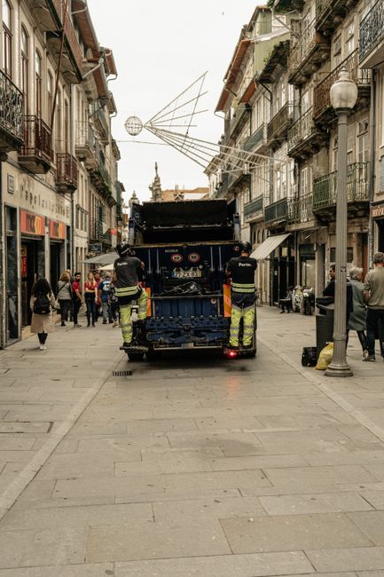 A street scene in an urban area featuring a blue rubbish collection truck operated by Rubbish Clearance Paddington positioned in the middle of the paved pedestrian street. The truck's rear is open, revealing various waste collection compartments and tools, with two workers in high-visibility uniforms standing at the back, engaging with the vehicle. Surrounding the truck, several pedestrians walk along the street, some observing the waste collection process, while others are engaged in conversation or browsing store fronts on the ground level of the multi-storey buildings on either side. These buildings exhibit traditional European architectural details, including balconies with metal railings and stone facades, some with awnings or potted plants. A street lamp with a rounded lantern is visible to the right of the truck, and festive decorations, resembling geometric wireframe structures and a star, hang overhead against a pale sky. The overall scene captures a typical city environment involving periodic waste management activities, with an emphasis on private refuse collection services in a busy shopping district.