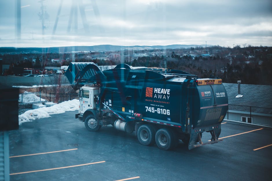 A black waste collection truck operated by Heave Away is parked in an outdoor parking lot during daylight hours, with snow piled along the edges of the asphalt. The truck features white and orange branding and contact information on its side, including the text 'Heave Away Waste & Environmental' and a phone number. The truck's rear compartment, used for hauling rubbish, is closed in this image. In the background, there are residential buildings with dark roofs, and a hilly landscape under an overcast sky, indicating cold winter weather. The parking area has visible parking lines and a slight reflection on the truck's shiny exterior, suggesting clean and well-maintained equipment suitable for private waste disposal services, often used for on-site clearance or commercial rubbish removal. The overall scene emphasizes professional waste collection activities in an environment where independent disposal methods are prevalent, aligning with services such as those offered by Rubbish Clearance Paddington for local businesses.