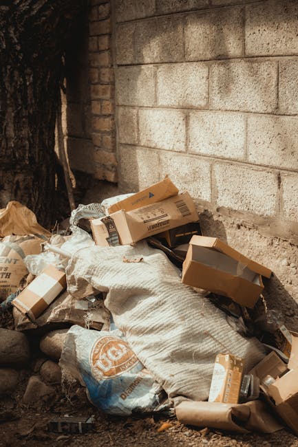 A pile of discarded cardboard boxes, some flattened and others partially intact, along with plastic packaging and paper waste, are gathered at the base of a brick and concrete wall on a dirt surface. The boxes are brown with printed labels and barcodes, and some are torn or crumpled. A large woven fabric sack is also part of the debris, with the waste appearing to be accumulated in an outdoor area, possibly near a building or garden boundary. The environment suggests a collection point for household or commercial rubbish awaiting removal, managed by a rubbish clearance service. The surrounding area includes visible soil and small rocks, with a section of a tree trunk to the left, indicating an exterior setting. The lighting is natural, casting soft shadows, and the scene captures an example of private waste accumulation that could be handled through an independent rubbish disposal or clearance service.