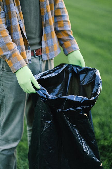 A person wearing a yellow and gray plaid shirt with a green long-sleeve underneath and gray trousers stands outdoors on a grassy area, holding open a large black trash bag lined with a reflective silver interior. The person is wearing light green gloves, and the trash bag appears to be partly filled with waste. The background features a blurred grassy landscape, suggesting an outdoor environment suitable for rubbish collection or waste disposal activities. This scene is representative of waste management tasks, such as rubbish removal or site clearance, potentially supported by private waste disposal services like Rubbish Clearance Paddington, highlighting the process of handling rubbish in a professional and organized manner.
