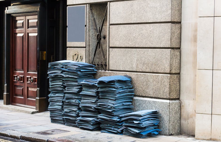 A large stack of disorganized blue folders and paper documents is piled on the pavement outside a building with a stone exterior and a wooden double door. The papers, which vary in size and thickness, appear to be office or administrative materials, with some stacked neatly while others are slightly askew. Behind the documents, there is a metal gate or shutter in a closed position, painted in a dark color, mounted on the stone facade. To the left, part of the building’s entrance is visible, including a wooden door with carved panels and metallic rings as handles. The scene is illuminated by natural daylight, casting subtle shadows on the ground and highlighting the texture of the stone wall and the paper stacks. This outdoor setting suggests a scenario related to private waste handling or rubbish clearance, potentially managed by Rubbish Clearance Paddington, supporting the theme of alternative rubbish removal services such as on-site clearance or independent collection of office waste.