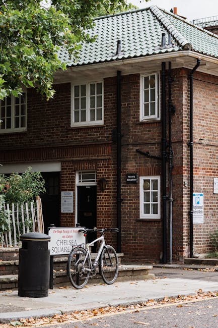 The image shows a two-storey brick building with a grey tiled pitched roof in an urban setting, partially obscured by leafy green tree branches in the foreground. The building features white-framed double-glazed windows on the upper and lower floors, with a small window on the side and a larger window at the front. A black downpipe runs vertically along the corner of the building, connected to the gutter system. In front of the building, on the paved sidewalk, there is a bicycle parked upright, secured to a small metal stand. To the left of the bicycle is a black cylindrical trash bin, and behind it, a white and red sign propped against a low fence indicates an area associated with Rubbish Clearance Paddington. The environment suggests a quiet residential street, with some fallen leaves scattered on the pavement and a blue parking restriction sign attached to the building's wall. The scene appears to be during daylight with natural lighting, capturing a typical urban rubbish collection or clearance setting that might be relevant to independent waste disposal services in Paddington.