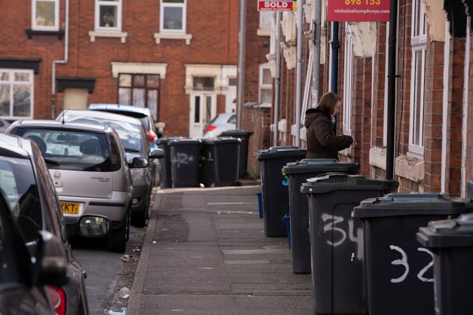 A woman standing on a residential street pavement, positioned next to a series of dark gray wheelie bins with white painted numbers, likely for rubbish collection or private waste disposal. The bins are lined up along the front of red-brick terraced houses, with some lids open or slightly tilted. The sidewalk is narrow, with a few pieces of litter scattered near the curb, and the street has several parked vehicles including a small silver hatchback in the foreground and other cars further along. The background features more terraced houses with white window frames and brick facades, and a red sign partially visible on the right indicates police or security service. The environment suggests a typical urban area where private waste management services like those offered by Rubbish Clearance Paddington could be relevant. The scene is captured in natural daylight, with ambient light illuminating the scene evenly, highlighting the textures of the brickwork, vehicle surfaces, and the plastic of the waste bins, supporting the context of community-level refuse management and alternative rubbish clearance services.