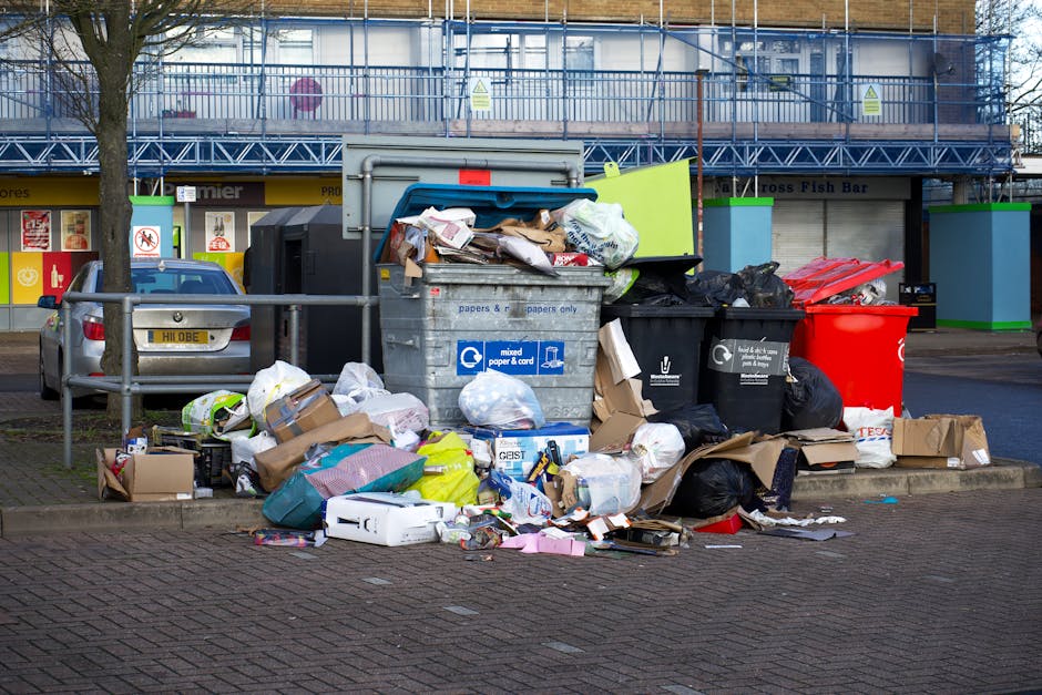 An outdoor scene showing a large overflowing collection of mixed waste and rubbish bags placed on a paved sidewalk beside a low metal railing. The waste includes cardboard boxes, plastic bags, paper, and various discarded packaging materials. A prominent grey waste container with a blue label reading 'mixed paper & card' is partially open, with papers and cardboard protruding from its top. To the right, several black and red rubbish bins are present, some filled with waste, and one bin appears to be damaged or overflowing. Behind the rubbish, there is a parking area with a small silver car parked adjacent to the curb, and in the background, a building with commercial storefronts under a blue framework and scaffolding, indicating recent construction or refurbishment. The scene is illuminated by natural daylight, contrasting the disorderly pile against the urban environment, reflecting a typical scenario where private waste collection services, such as those offered by Rubbish Clearance Paddington, might be engaged to manage excess rubbish beyond standard communal bin capacity.