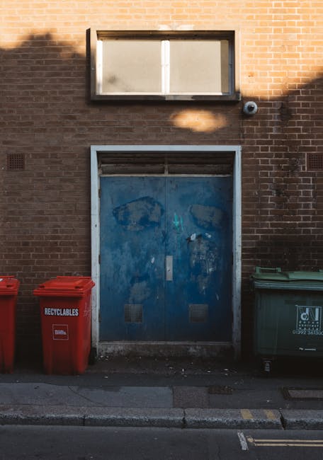 A woman standing on a residential street pavement, positioned next to a series of dark gray wheelie bins with white painted numbers, likely for rubbish collection or private waste disposal. The bins are lined up along the front of red-brick terraced houses, with some lids open or slightly tilted. The sidewalk is narrow, with a few pieces of litter scattered near the curb, and the street has several parked vehicles including a small silver hatchback in the foreground and other cars further along. The background features more terraced houses with white window frames and brick facades, and a red sign partially visible on the right indicates police or security service. The environment suggests a typical urban area where private waste management services like those offered by Rubbish Clearance Paddington could be relevant. The scene is captured in natural daylight, with ambient light illuminating the scene evenly, highlighting the textures of the brickwork, vehicle surfaces, and the plastic of the waste bins, supporting the context of community-level refuse management and alternative rubbish clearance services.