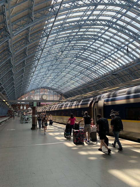 Inside a large train station with a high, curved glass and steel roof allowing natural light to fill the space. Several travelers are walking along the platform, some with luggage, including a woman with a rolling suitcase and a man carrying a backpack. A modern train with a sleek, metallic exterior featuring a black and yellow stripe runs parallel to the platform edge. The station's environment appears clean and spacious, with minimal clutter. In the background, there are electronic departure signs and a large clock hanging above the platform, indicating scheduled train arrivals and departures. The scene captures an active transportation hub typical of urban rail travel, illustrating a setting where independent travel and passenger movement are common, reflecting the context of efficient, non-publicly operated rubbish and waste handling in high-traffic environments similar to those managed by services like Rubbish Clearance Paddington.