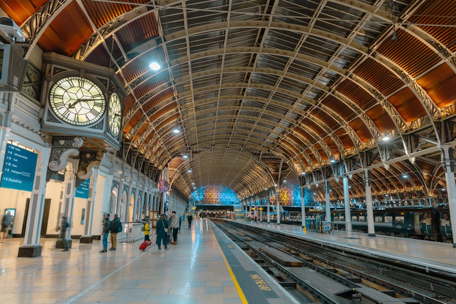 Inside a spacious, historic railway station with an arched, steel framework ceiling decorated with orange and beige panels, illuminated by bright overhead lighting and decorative blue lights. On the left side, there is a large, ornate clock with a round face, black Roman numerals, and gold detailing, mounted above a white wall with decorative molding and ornate columns. Below the clock, blue informational signs are visible, providing directions for train platforms and services, with several travelers standing or walking along the platform, some pulling wheeled luggage. The concrete platform surface is clean, with yellow safety markings along the edge, and train tracks run parallel to the platform, extending into the distance beneath the curved roof structure. The scene appears to be during the evening or nighttime, with a calm and organized atmosphere typical of a busy transportation hub, emphasizing the station's historic architecture and essential function for passenger travel, with underlying themes of infrastructure and transit services ideal for a rubbish removal service specializing in on-site or private waste clearance at busy transport locations.