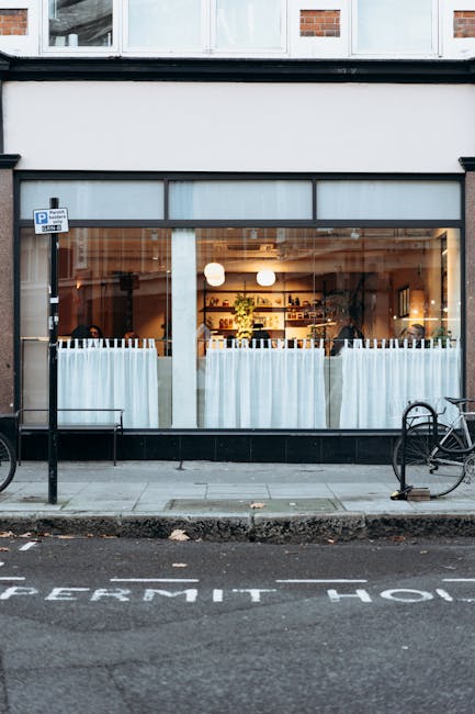 The image depicts a large storefront window of a modern café or restaurant positioned at street level, with sheer white curtains partially drawn across the lower part of the glass. Inside, warm lighting illuminates a tidy interior featuring wooden shelves stocked with various items, possibly beverages or kitchenware, and a few potted plants adding greenery to the scene. The exterior consists of a black-framed glass display and adjacent brickwork sections, with a narrow sidewalk in front. A small bicycle is chained to a black metal bicycle stand on the right side of the image, while on the left, another bike is parked, both facing the street. A designated parking bay with a “permit only” marking is visible on the road, along with a small parking sign attached to a pole near the storefront. The setting appears to be in an urban area, with muted colors and natural light creating a neutral, professional atmosphere suited to the context of independent waste disposal or rubbish management, where private collection services such as those provided by Rubbish Clearance Paddington could be relevant to ensuring tidy premises or waste removal outside the scope of local authority services.