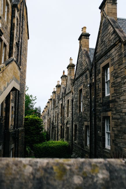 A narrow alley between historic stone buildings with dark, weathered brick facades, featuring multiple chimney stacks on pitched slate roofs. The buildings have small, rectangular white-framed sash windows, some with visible glass panes. The scene is viewed from a slightly elevated position, with a moss-covered stone wall or ledge in the foreground. Greenery, including a bush or small tree, is visible at the bottom of the alley, suggesting an urban residential area. Overcast sky provides diffuse lighting, highlighting the textures of the stone walls. The setting evokes a traditional UK terrace, where private waste collection or on-site clearance could be relevant for maintaining cleanliness and managing rubbish efficiently, aligning with services offered by Rubbish Clearance Paddington in a historic Paddington terrace context.
