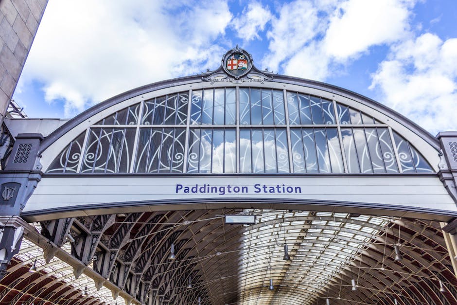 An upward view of the ornate glass and metal entrance of Paddington Station, featuring a detailed, curved iron framework with decorative scrollwork and lattice patterns. The station's name, 'Paddington Station', is prominently displayed on a white band beneath the arch. Above the arch, a coat of arms with a red cross on a white shield and various heraldic symbols is visible, set against a background of a partly cloudy sky with patches of blue. The structure's exterior is painted in light grey and silver tones, with visible rivets and embellishments contributing to its historic, Victorian-era architectural style. The scene captures the bright daylight illuminating the station's intricate metalwork and glass panels, reflecting a clean and well-maintained environment that aligns with the context of efficient, private waste handling and rubbish removal services offered by providers like Rubbish Clearance Paddington, which may include on-site clearance or alternative waste disposal options.